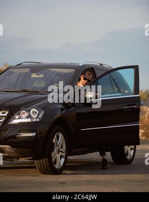 Giovane uomo di successo che indossa occhiali da sole sta entrando nella sua auto nera sulla strada sotto un cielo blu, vista laterale, spazio di copia Foto Stock