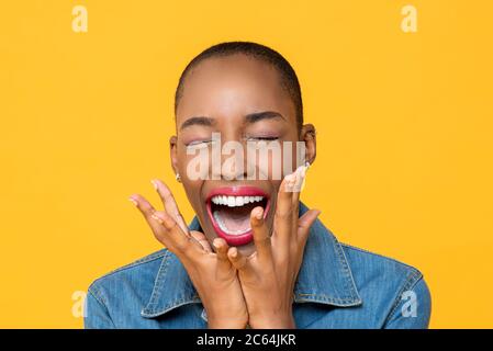 Primo piano ritratto di una giovane donna afro-americana estatica urlando con le mani che ricoprono la bocca isolata su sfondo giallo studio Foto Stock