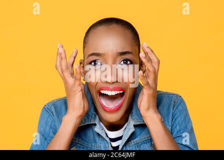 Primo piano ritratto di giovane donna afro-americana spaventata urlando con entrambe le mani sollevate in uno studio isolato sfondo giallo Foto Stock