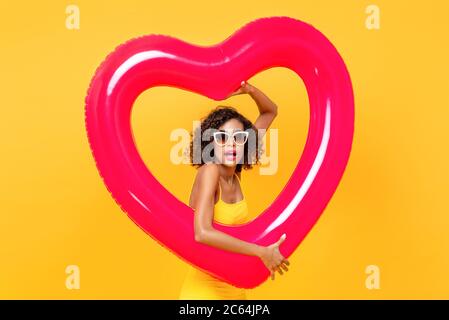 Ritratto di donna afro-americana sorpresa indossare occhiali da sole tenendo il pallone forma cuore in studio isolato sfondo giallo Foto Stock