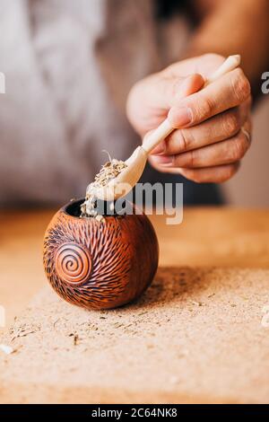 Cucina di accoppiamento - chiusura di una mano femminile spruzza le foglie di mate secche in un calabash con un cucchiaio di legno Foto Stock