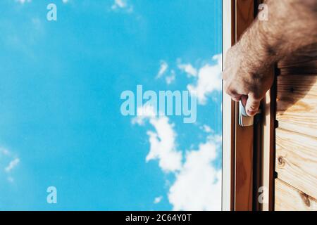 Una mano dell'uomo apre l'otturatore della finestra tramite la maniglia - una finestra a doppio vetro nella mansarda con una vista del cielo blu Foto Stock