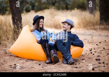 Giovane coppia felice in cappello passare il tempo insieme riposarsi in un divano gonfiabile arancione nella foresta autunnale Foto Stock