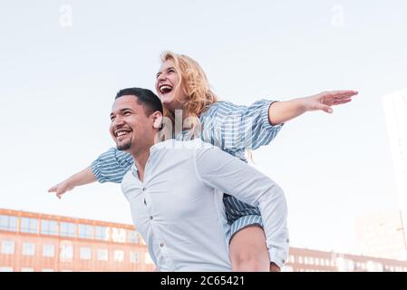 una coppia gioca felicemente, si apre sulle spalle le braccia e finge di volare, la porta con gioia, sorrisi felici e ottimisti di una co multietnica Foto Stock