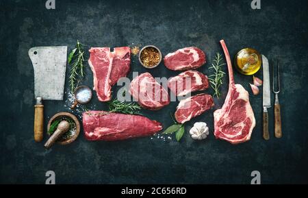Varietà di bistecche di carne di manzo cruda per grigliare con condimento e utensili su tavola rustica scura Foto Stock