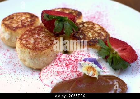 Frittelle al formaggio di cottage, scirniki con frutti di bosco freschi per la colazione su piatto bianco, sfondo tavola di legno. Frittelle dolci russe con fragole e. Foto Stock