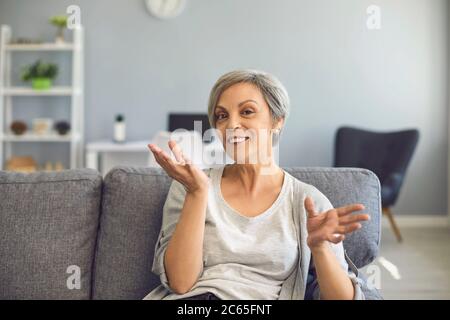 Donna anziana felice con i capelli grigi dice guardando la macchina fotografica mentre si siede su un divano a casa. Foto Stock