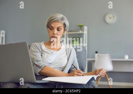 Lavorare online a casa. Una donna anziana con capelli grigi lavora a distanza utilizzando un computer portatile mentre si siede in una sedia nella stanza Foto Stock