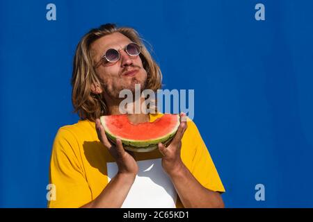 Giovane attraente con lunghi capelli biondi che mangia mezzo anguria e che si gode su uno sfondo blu chiaro. Concept estate, sole, calore, frutta, vacanze Foto Stock