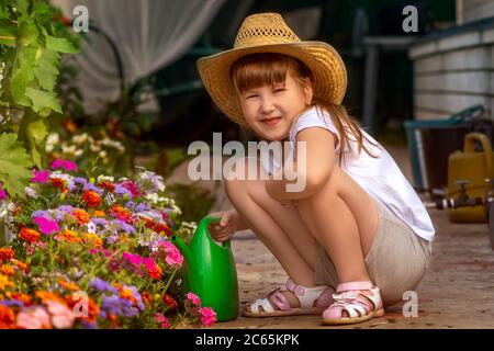 Bella ragazza sorridente in paglia cowboy cappello all'aperto in giardino si prende cura di letto di fiori da annaffiarlo da annaffiatura verde lattina. Foto Stock