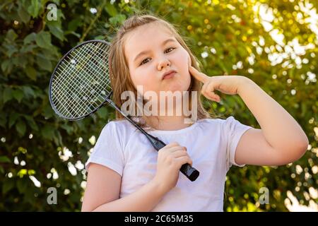 Una bambina scherzosamente rende un viso glamour. Carina bambina che gioca badminton all'aperto in una calda e soleggiata giornata estiva. Foto Stock