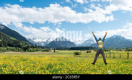 Hiker felice riuscito con il salto dello zaino con le braccia sollevate sul prato del fiore del dente di leone. Montagne innevate e casa tradizionale. Baviera, Alpi Foto Stock