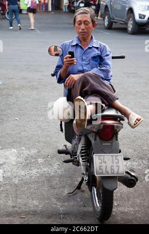 Uomo vietnamita in camicia blu che si rilassa sulla sua moto sul lato della strada con gambe incrociate messaggi sul suo cellulare. Foto Stock