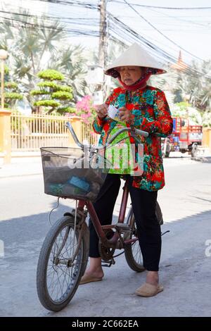 Donna vietnamita locale seduta sulla sua bicicletta con un cimpaniere sul marciapiede mentre fuori shopping controllare alcuni biglietti in strada vicino a My Tho City, Vietnam Foto Stock