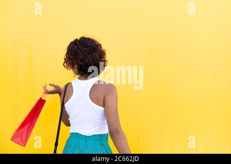 Vista posteriore di una donna che cammina con una borsa accanto a un muro giallo. Spazio per il testo. Concetto di shopping, regali e vendite. Foto Stock