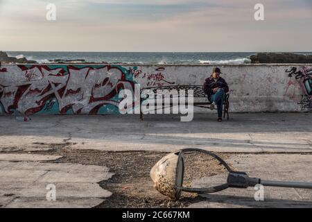 Vecchio seduto su una panchina di ferro di fronte al muro di banchina a Essaouira Foto Stock