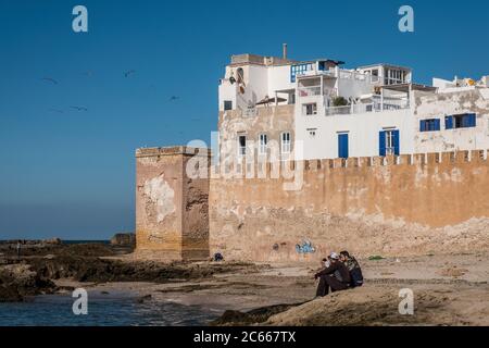 Uomini seduti su rocce nel mare di fronte alle mura della città di Essaouira Foto Stock