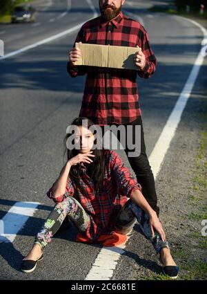 Coppia in amore che viaggia da hitchtrekking, ragazza siede sulla strada, spazio copia. L'uomo tiene il cartello di cartone, prova a fermare l'auto. Concetto di viaggio e di escursionismo. Coppia con volti pensivi viaggia con auto stop. Foto Stock