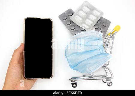 Mini carrello con maschera medica protettiva piena di pillole accanto alla mano donna con telefono cellulare con schermo verde mockup. Sfondo bianco. interno Foto Stock