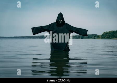 un uomo in un accappatoio nero si trova nel lago con le mani allungate e guarda l'acqua sullo sfondo della natura al giorno Foto Stock