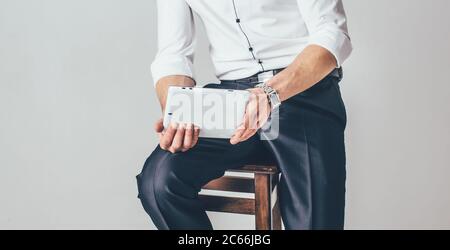 l'uomo tiene una tavoletta in mano sullo sfondo bianco. Si siede su una sedia vestita con una camicia bianca e pantaloni Foto Stock