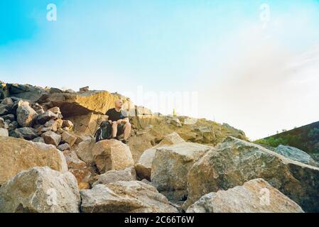 un viaggiatore si siede sulla cima di una roccia sulla pietra sullo sfondo di cielo senza nuvole. Un turista guarda alla distanza dalla bellezza della natura Foto Stock