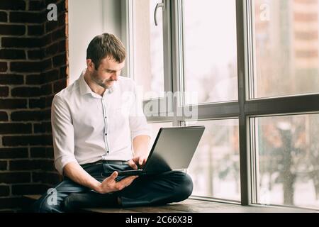 uomo d'affari si siede sul davanzale della finestra in posa di lotus e lavora sul laptop nel suo armadietto Foto Stock