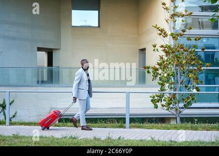 Afroamericano in tuta bianca e valigia rossa. Uomo d'affari. Uomo d'affari in viaggio. Uomo in viaggio. Foto Stock