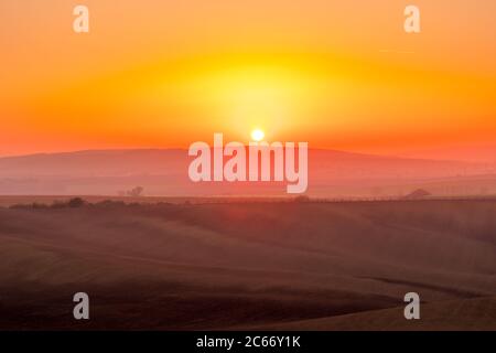 Il sole catturato durante l'ultimo momento prima di andare oltre l'orizzonte con vista di un campo coperto di nebbia e le fattorie e i campi ondulati in t Foto Stock