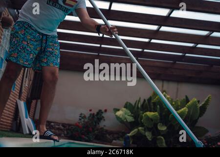 Persona che pulisce e clorurando la piscina in un caldo pomeriggio estivo, sta facendo funzionare il pulitore e aggiungendo polvere di cloro Foto Stock