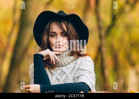 una ragazza sorprendente con cappello nero e maglione a maglia tiene la mano sul mento e guarda la macchina fotografica sullo sfondo del parco in autunno Foto Stock