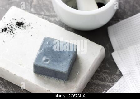 Sapone fatto a mano con carbone attivo su marmo, polvere di carbonio e un asciugamano di cotone su fondo grigio. Foto Stock