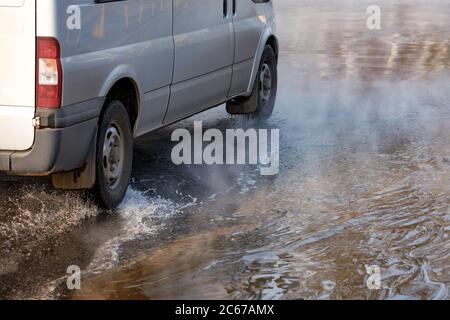 La carreggiata è piena di acqua calda dopo la svolta della rete di riscaldamento della città, l'auto passa attraverso le pozzanghere in una nuvola di vapore caldo. Foto Stock