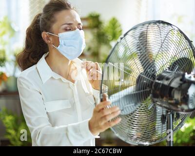 Estate calore. Triste giovane donna in blusa bianca con ventilatore elettrico sensazione di calore estivo nel moderno soggiorno in sole calda giornata estiva. Foto Stock