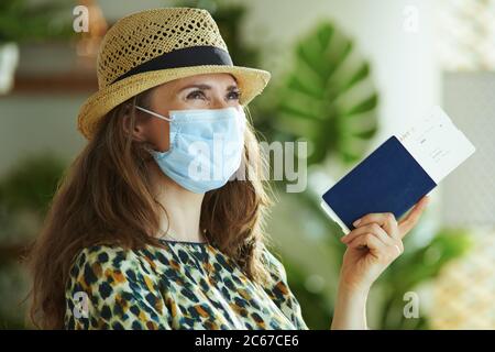 Vita durante la pandemia di covid-19. Donna trendy sognante in cappello di paglia con maschera medica, passaporto e biglietto aereo che guarda in lontananza. Foto Stock