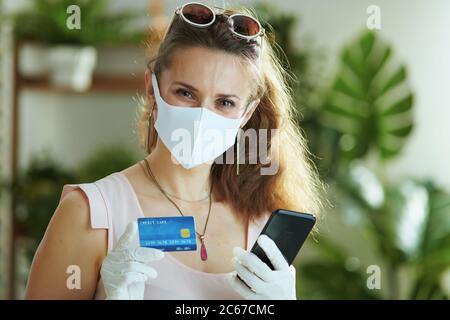 Vita durante la pandemia di covid-19. Moderna donna di mezza età acquirente in blusa rosa con maschera medica, guanti di gomma bianca, carta di credito e smartphone maki Foto Stock