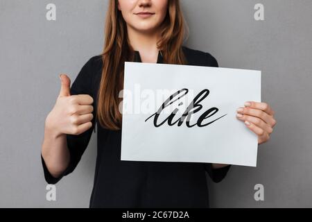 Primo piano foto di mani di ragazza che tengono la cartolina e mostrando il pollice grande su sfondo grigio Foto Stock