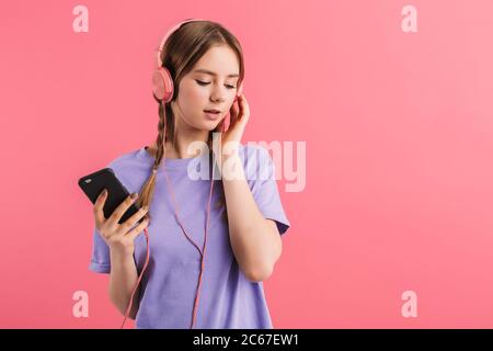 Giovane ragazza attraente con due trecce in t-shirt lilla ascolto di musica in cuffia con cellulare in mano mentre sognando da parte su rosa Foto Stock