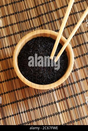 Concetto di cibo sano. Riso nero selvatico con bastoncini di chopsticks in una ciotola di legno su un tappeto di bambù. Messa a fuoco selettiva. Vista dall'alto. Foto Stock