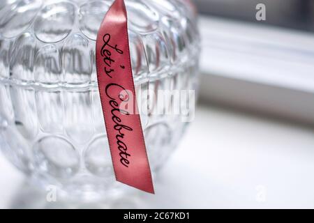 Contenitore decorativo in vetro retrò con motivo a nastro rosa con testo festeggiamo su un davanzale bianco. Foto Stock
