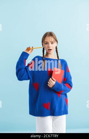 Giovane ragazza attraente con due trecce in pullover oversize con cuori rossi che tengono il notebook e la matita in mani mentre si guarda stupito in macchina fotografica sopra Foto Stock