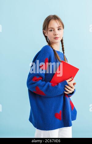 Giovane bella ragazza con due trecce in pullover oversize con cuori rossi che tengono il notebook in mano mentre sognando in macchina fotografica su blu Foto Stock