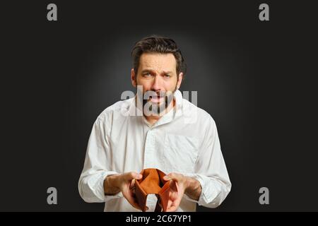 Uomo caucasico sconvolto che tiene in mano un portafoglio vuoto. Il concetto di emozioni e crisi finanziaria. Ritratto isolato su sfondo grigio. Foto Stock