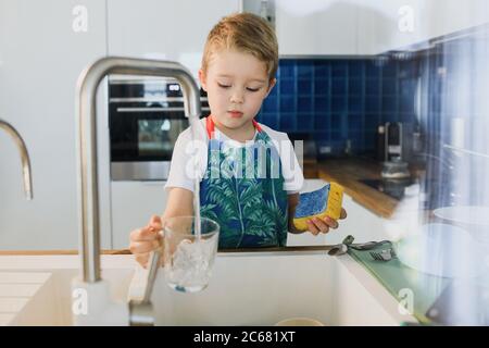 un ragazzo in grembiule lava i piatti in cucina a casa Foto Stock