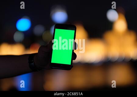 una silhouette a mano che mostra lo schermo verde dello smartphone nelle ore notturne. Bella sfocatura città dorata luci di sfondo Foto Stock