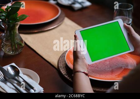 sulla spalla delle persone che usano un tablet con schermo verde nel ristorante. piatti sfocati sul tavolo da pranzo. Foto Stock