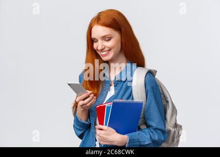 Bella bella femmina con capelli rossi, andare al college, tenendo zaino, notebook sorridenti come risposta messaggio amico, chiacchierare guardando mobile Foto Stock