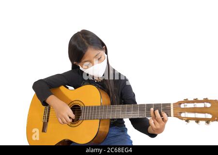 Una studentessa asiatica che indossa una maschera e suona la chitarra a casa. L'immagine mostra l'idea durante la quarantena domestica per ridurre la diffusione del coron Foto Stock