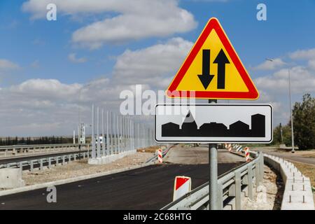 Cartello stradale temporaneo a due vie e il traffico segno l'inizio del villaggio contro il cielo e la strada in costruzione. Foto Stock