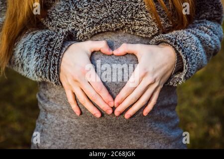uomo che tiene il ventre della moglie incinta che fa un cuore. Donna incinta e marito amorevole abbracciano il ventre a casa. Cuore dalle mani di un multi Foto Stock